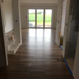 Nowo ułożone panele podłogowe w odcieniach brązu w jasnym wnętrzu z widokiem na zewnątrz przez przeszklone drzwi przesuwne; widoczne butelki z klejem na schodach i niebieska taśma zabezpieczająca...