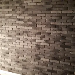 Ściana z szarych cegieł dekoracyjnych, nierównomierne oświetlenie podkreśla fakturę, widoczna rurka pionowa w prawym dolnym rogu.