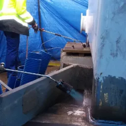 Pracownik w żółtej kurtce, kasku i rękawicach spawa element konstrukcyjny z użyciem palnika, widoczne iskry i niebieskie węże doprowadzające gaz, tło stanowi niebieska plandeka.