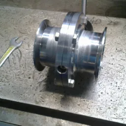 Element z polerowanej stali z nakrętkami na szorstkiej metalowej powierzchni warsztatowej, obok leży klucz płaski.