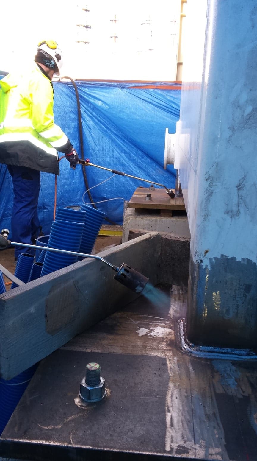 Pracownik w żółtej kurtce, kasku i rękawicach spawa element konstrukcyjny z użyciem palnika, widoczne iskry i niebieskie węże doprowadzające gaz, tło stanowi niebieska plandeka.