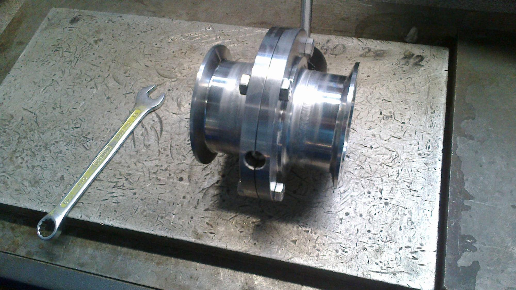 Element z polerowanej stali z nakrętkami na szorstkiej metalowej powierzchni warsztatowej, obok leży klucz płaski.