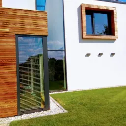 Nowoczesna fasada domu z drewnianą okładziną, dużymi oknami z ramami w kolorze antracytu i starannie utrzymanym trawnikiem.