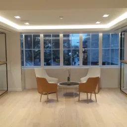 Nowoczesne wnętrze biurowe z podświetlanym sufitem, drewnianą podłogą, regałami z czarnego metalu i drewna, oraz strefą relaksu z krzesłami i stolikiem kawowym.