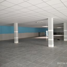 Polteam - Wnętrze dużego pomieszczenia z szarą podłogą z płytek, pomalowaną na niebiesko ścianą i białym sufitem kasetonowym, wspartego na szarych kolumnach z białymi pasami.