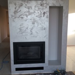 Polteam - Obudowa kominka z dekoracyjną szarą strukturą, z czarnym wkładem kominkowym i wnęką, zabezpieczona szarą włókniną i narzędziami na podłodze.