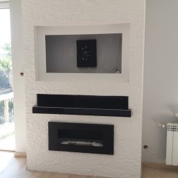 Polteam - Ściana z dekoracyjną białą okładziną, wnęką na telewizor z uchwytem, czarną półką i elektrycznym kominkiem z białymi kamyczkami w salonie z drewnianą podłogą.