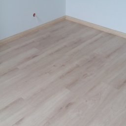 Polteam - Jasne panele podłogowe ułożone w rogu pomieszczenia, widoczne listwy przypodłogowe i fragment rolki zabezpieczającej podłogę.