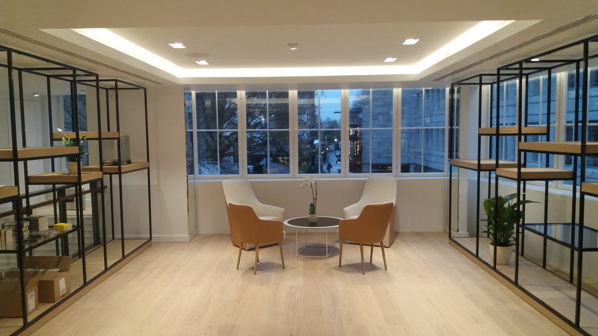 Nowoczesne wnętrze biurowe z podświetlanym sufitem, drewnianą podłogą, regałami z czarnego metalu i drewna, oraz strefą relaksu z krzesłami i stolikiem kawowym.