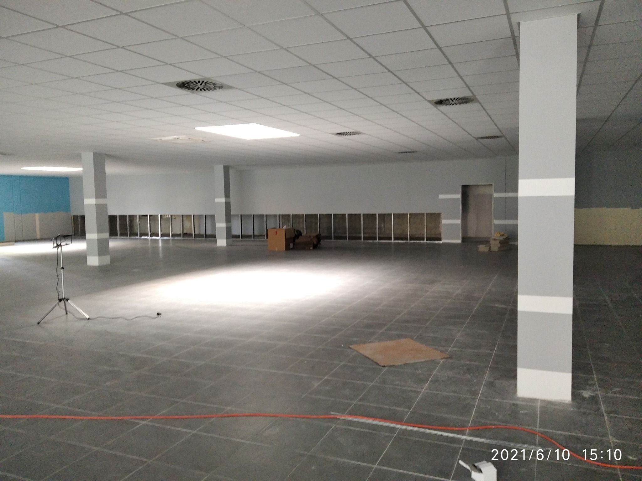 Przestronne wnętrze w trakcie prac wykończeniowych; widoczne szare kolumny z białymi pasami, podwieszany sufit z kratownicą, częściowo przygotowane ściany, oraz sprzęt oświetleniowy na statywie.