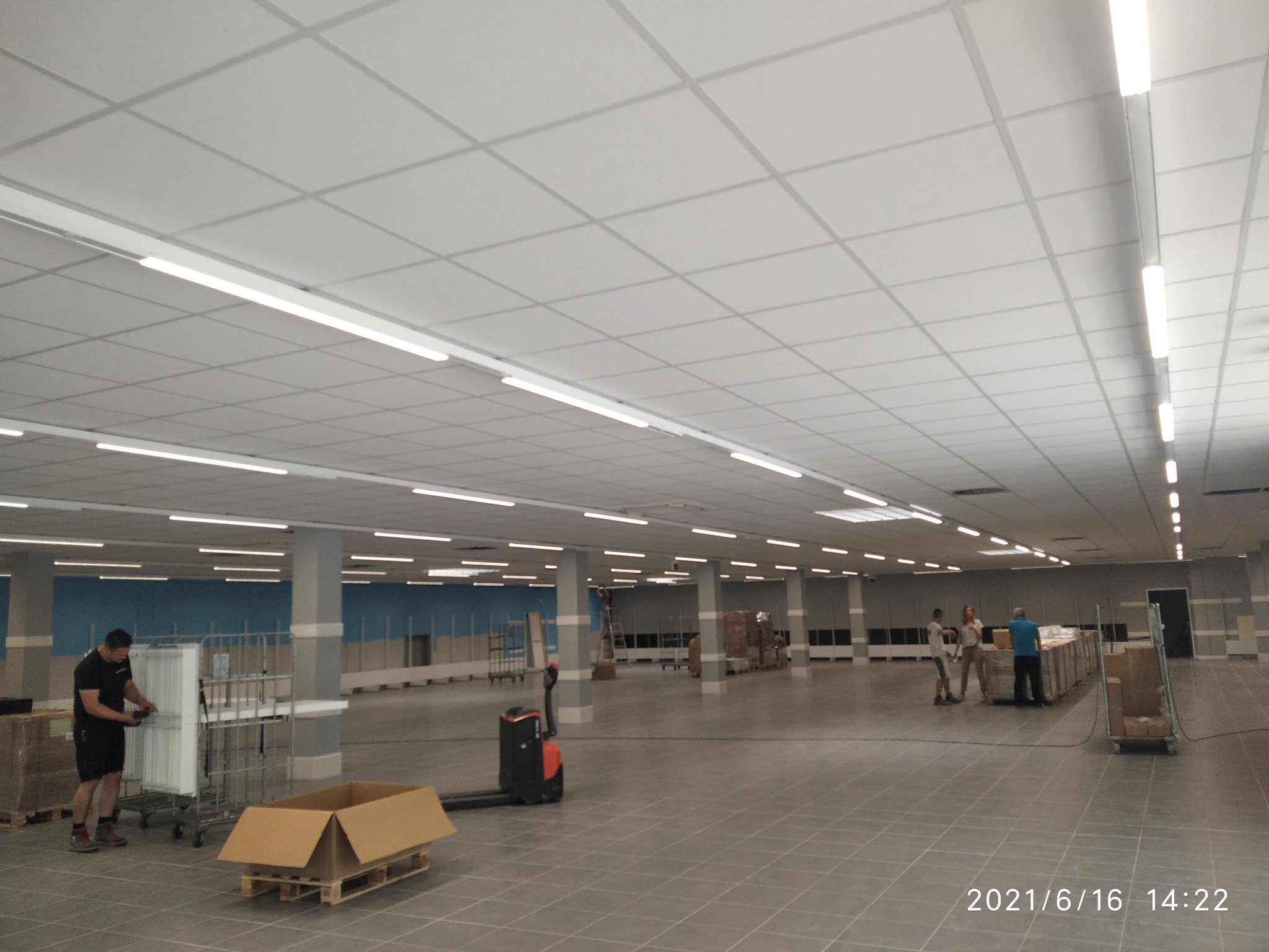 Przestronne wnętrze biurowe z podwieszanym sufitem kasetonowym i oświetleniem liniowym LED, pracownicy rozładowują materiały budowlane z palet i wózków transportowych.