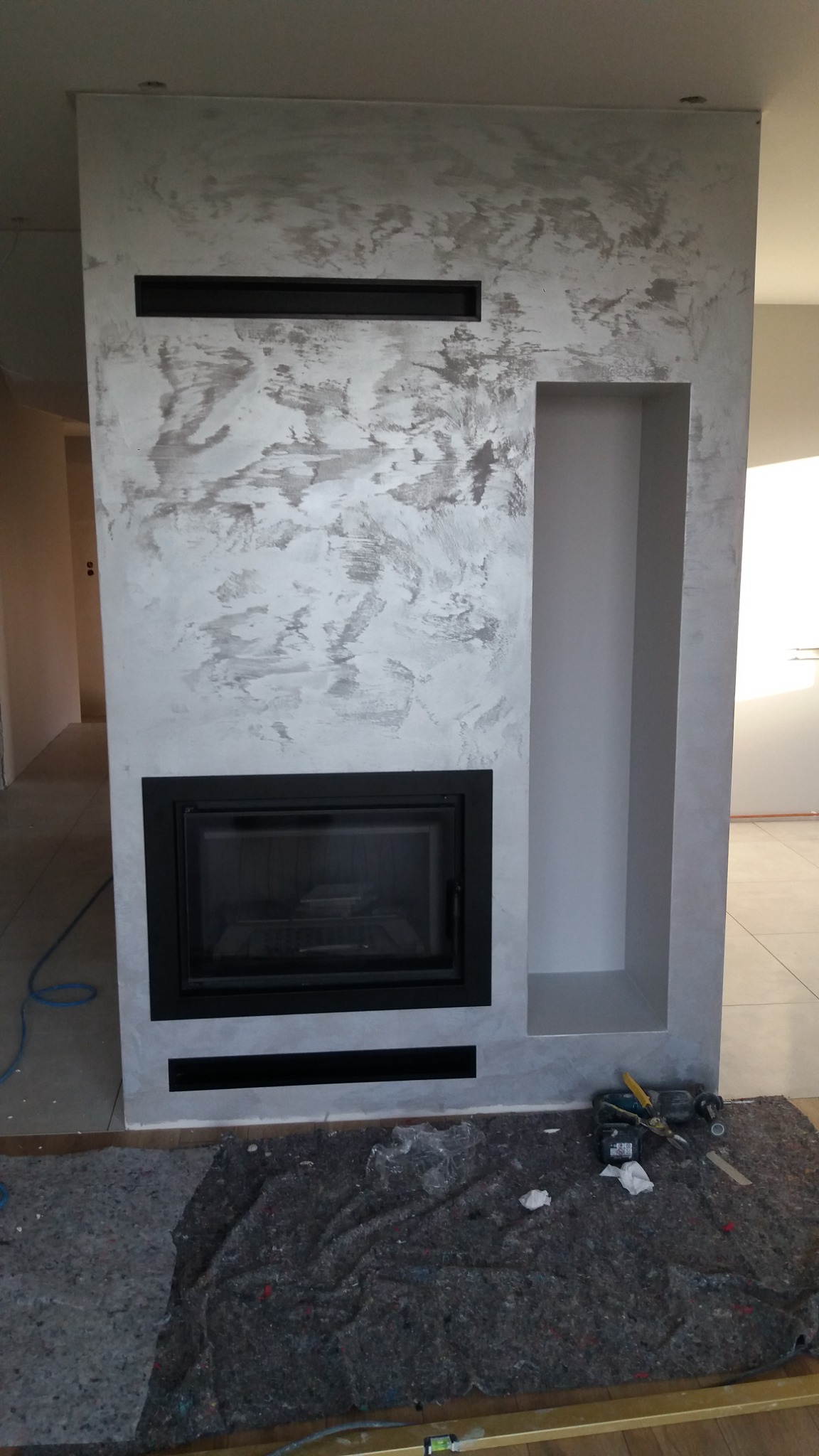 Obudowa kominka z dekoracyjną szarą strukturą, z czarnym wkładem kominkowym i wnęką, zabezpieczona szarą włókniną i narzędziami na podłodze.