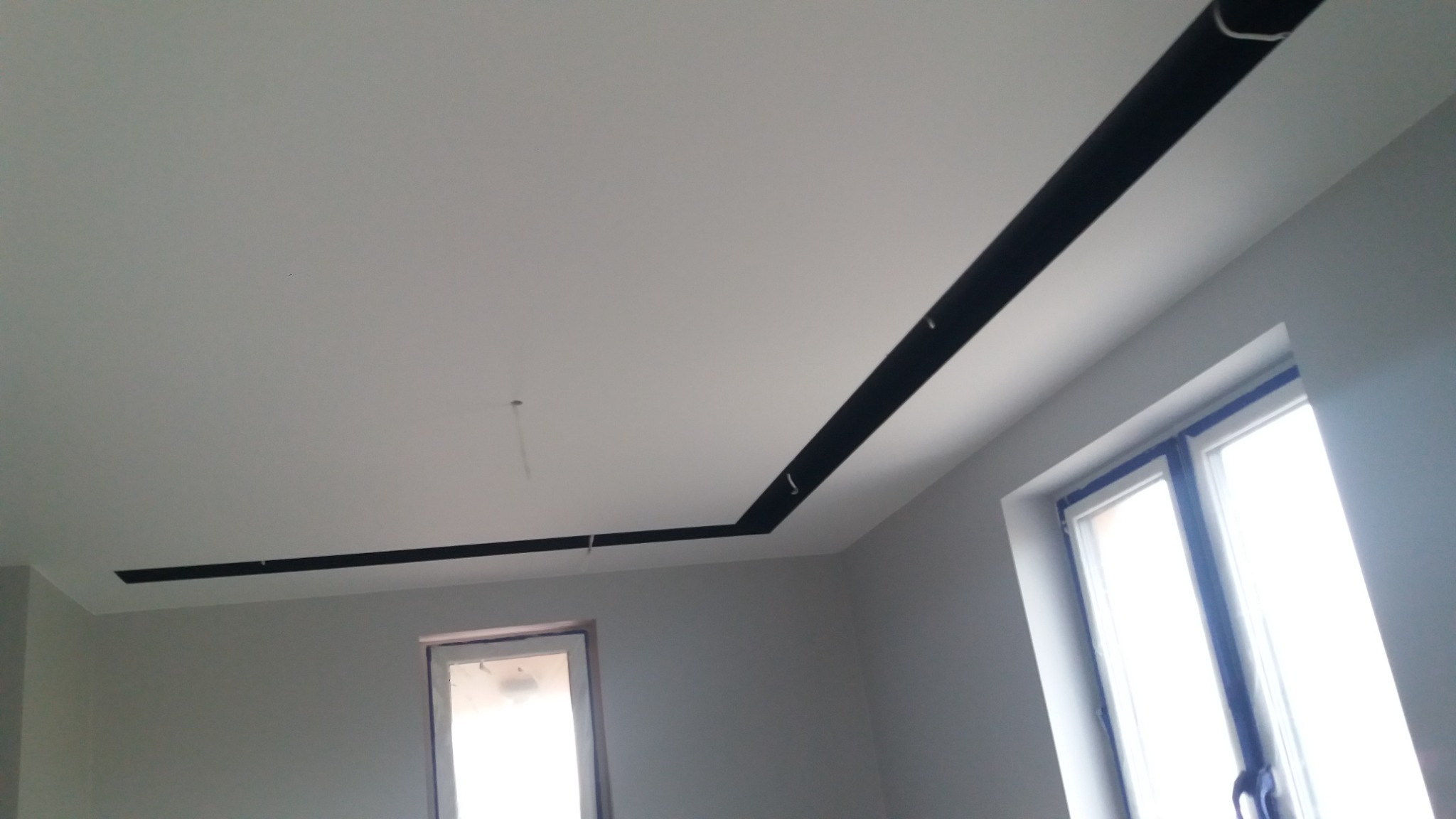 Świeżo pomalowane wnętrze z czarnym, dekoracyjnym obramowaniem sufitu, okna zabezpieczone taśmą malarską, widoczne niedoskonałości w wykończeniu.