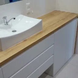 Nowoczesna łazienka z białą umywalką nablatową i szafką z drewnianym blatem, minimalistyczny design, białe płytki na ścianie, chromowana bateria.