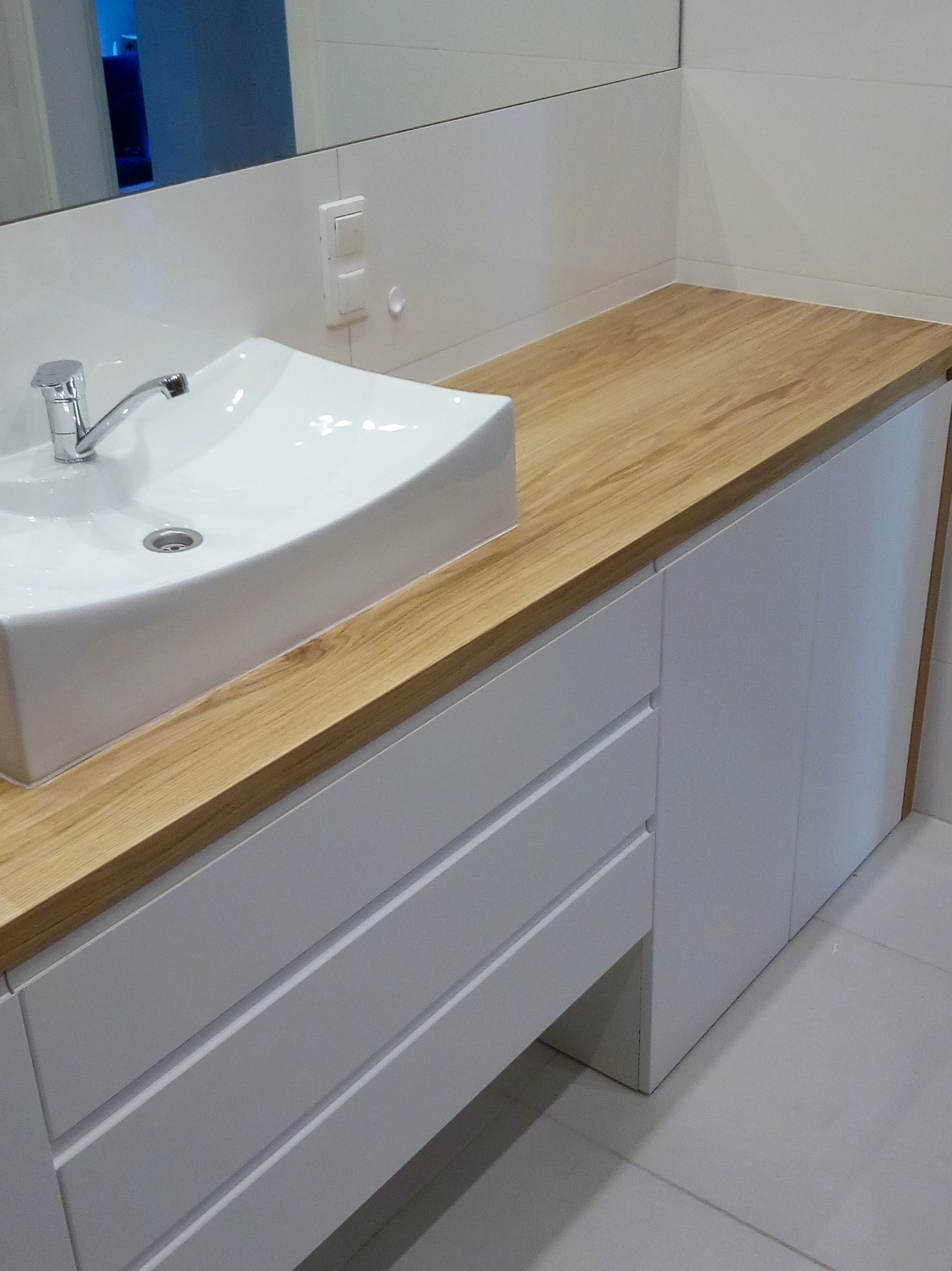 Nowoczesna łazienka z białą umywalką nablatową i szafką z drewnianym blatem, minimalistyczny design, białe płytki na ścianie, chromowana bateria.