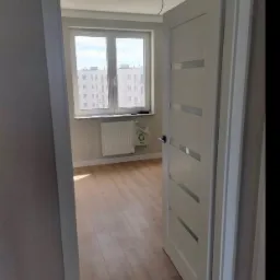 Widok na jasne, odświeżone wnętrze mieszkania z drewnianą podłogą, przez otwarte białe drzwi z poziomymi przeszkleniami, okno z widokiem na budynek mieszkalny w tle.