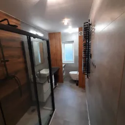 Nowoczesna łazienka z kabiną prysznicową typu walk-in z czarnymi ramami, umywalką nablatową na szafce, grzejnikiem drabinkowym w kolorze czarnym, toaletą i pralką w tle.