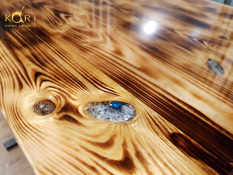 Blat biurka z jasnego drewna o wyraźnym usłojeniu, wykończony żywicą z zatopionymi kamieniami, widoczne logo firmy w lewym górnym rogu.