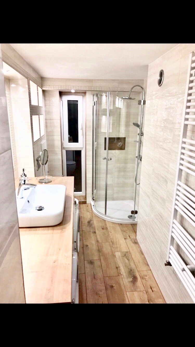 Wnętrze łazienki z kabiną prysznicową, umywalką nablatową na drewnianym blacie, grzejnikiem drabinkowym i podłogą z paneli imitujących drewno, widok na toaletę przez okno.