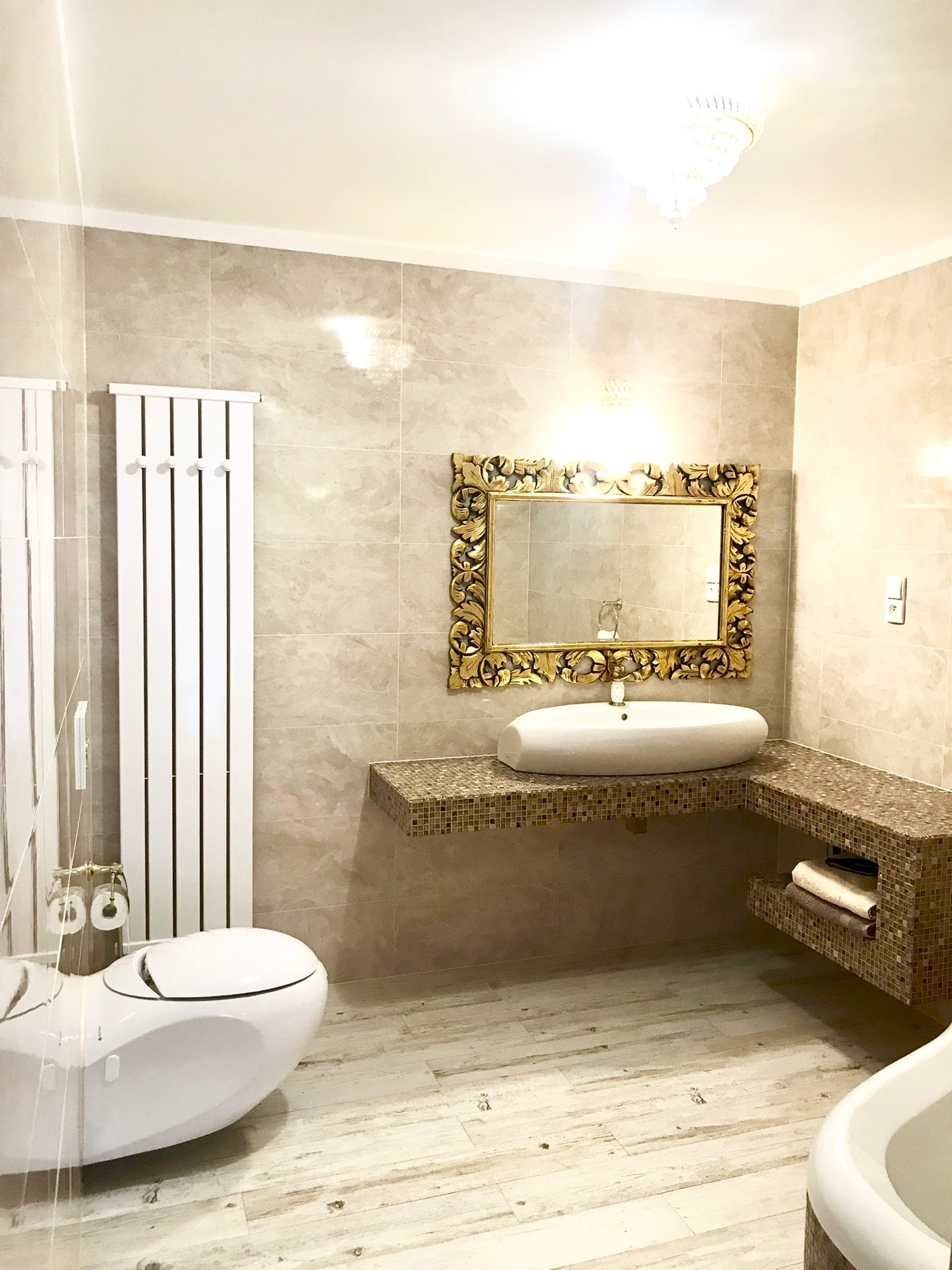 Elegancka łazienka w jasnych odcieniach z beżowymi płytkami, owalną umywalką na mozaikowym blacie, ozdobnym lustrem w złotej ramie i nowoczesną toaletą bez spłuczki.