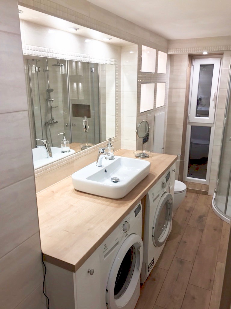 Jasna łazienka z pralkami zabudowanymi pod blatem z umywalką nablatową, kabina prysznicowa w lustrze, drewnopodobne panele na podłodze.