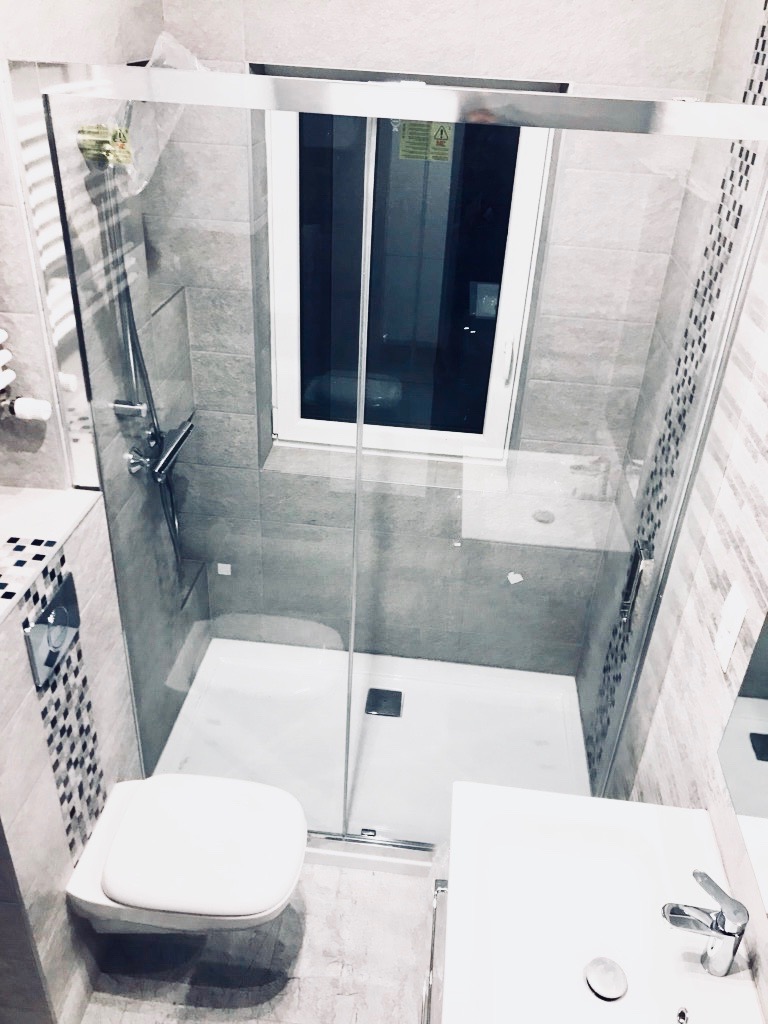 Nowoczesna łazienka z kabiną prysznicową typu walk-in, wiszącą toaletą i umywalką, wykończona szarymi płytkami z mozaikowymi akcentami, oświetlona przez okno z czarną ramą.