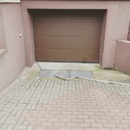 Brązowa segmentowa brama garażowa marki Hormann, z widocznym fragmentem uszczelki i odpływem liniowym przed nią, na tle elewacji w kolorze piaskowym.