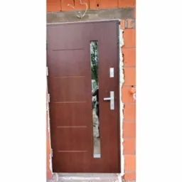 Brązowe drzwi wejściowe z podłużnym przeszkleniem, zamontowane w surowym murze z czerwonej cegły, widoczne przewody elektryczne nad ościeżnicą.