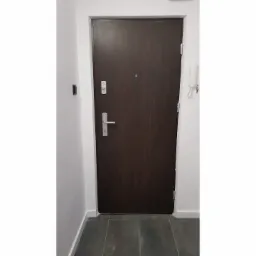 Ciemnobrązowe drzwi wejściowe do mieszkania z wizjerem i srebrną klamką, na ścianie biały domofon z kluczami.