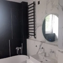 Nowoczesna łazienka z czarnymi i białymi marmurkowymi płytkami, okrągłym lustrem z podświetleniem LED, grzejnikiem drabinkowym i wolnostojącą wanną.