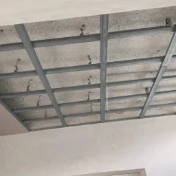 Metalowa konstrukcja sufitu podwieszanego z widoczną izolacją termiczną, częściowo pokryta płytą kartonowo-gipsową przy oknie.