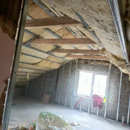 Poddasze w trakcie ocieplania wełną mineralną, widoczne metalowe profile konstrukcyjne, fragment muru z oknem, na podłodze resztki materiałów budowlanych oraz torba z wełną.