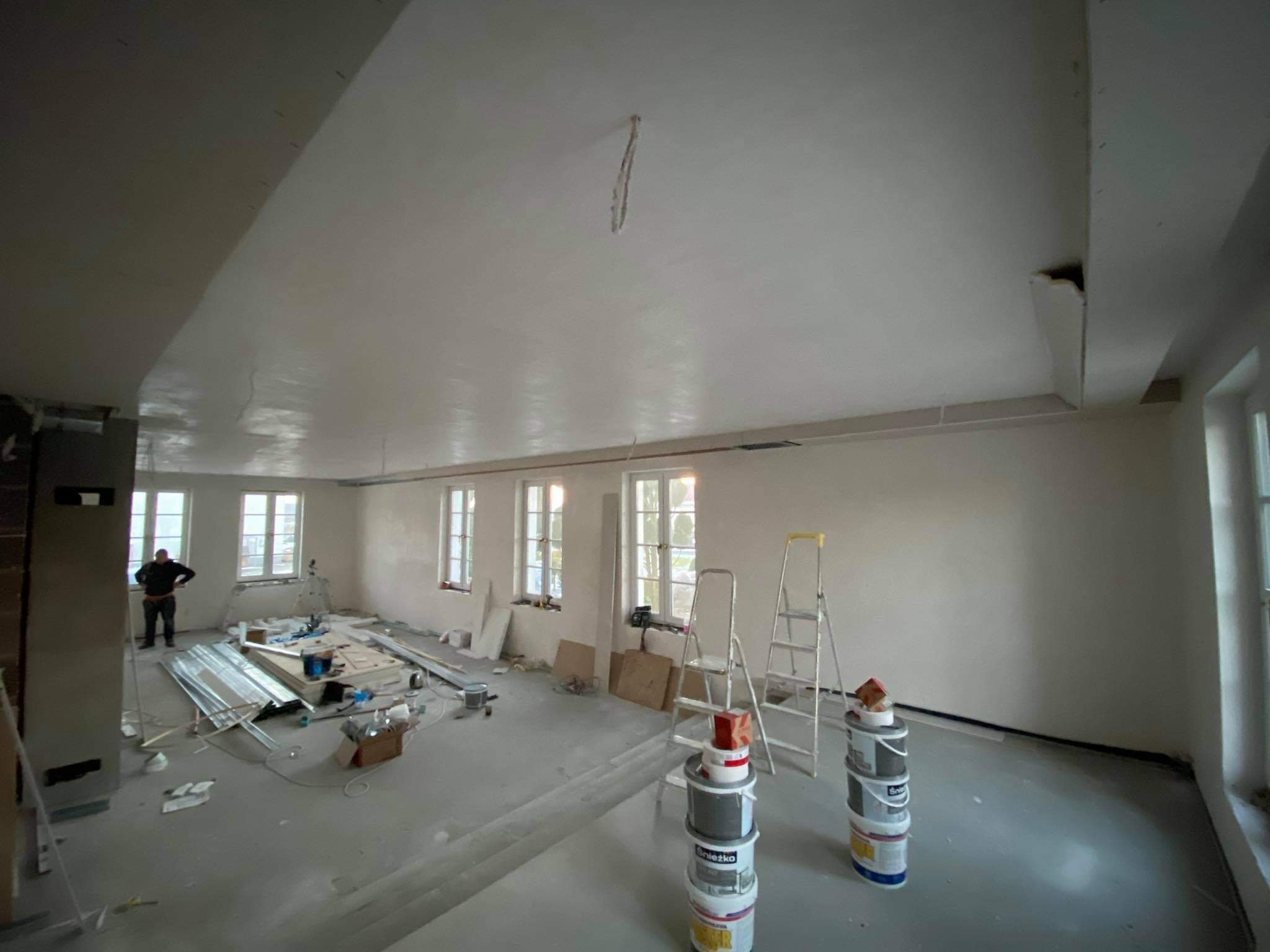 Wnętrze w trakcie prac wykończeniowych: świeżo wygładzona ściana i sufit, widoczne drabiny, farby, listwy przypodłogowe i narzędzia, osoba w tle.