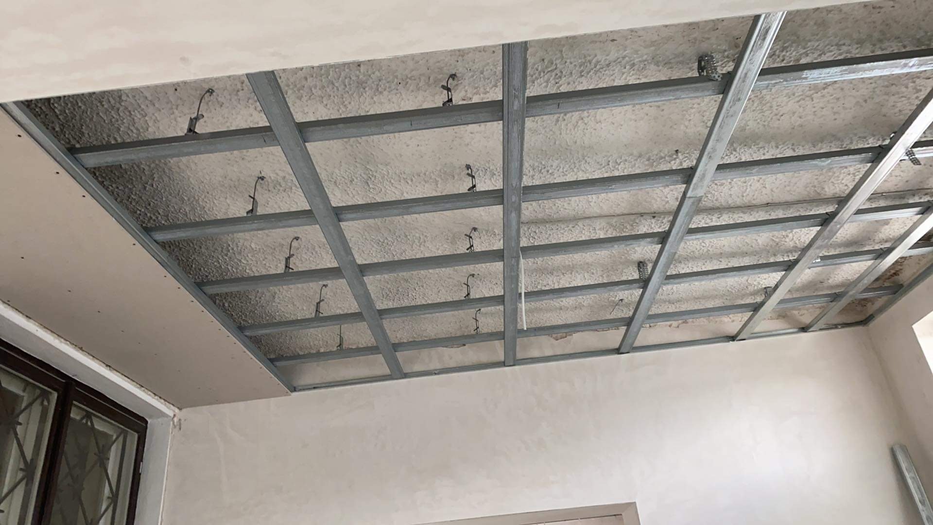 Metalowa konstrukcja sufitu podwieszanego z widoczną izolacją termiczną, częściowo pokryta płytą kartonowo-gipsową przy oknie.