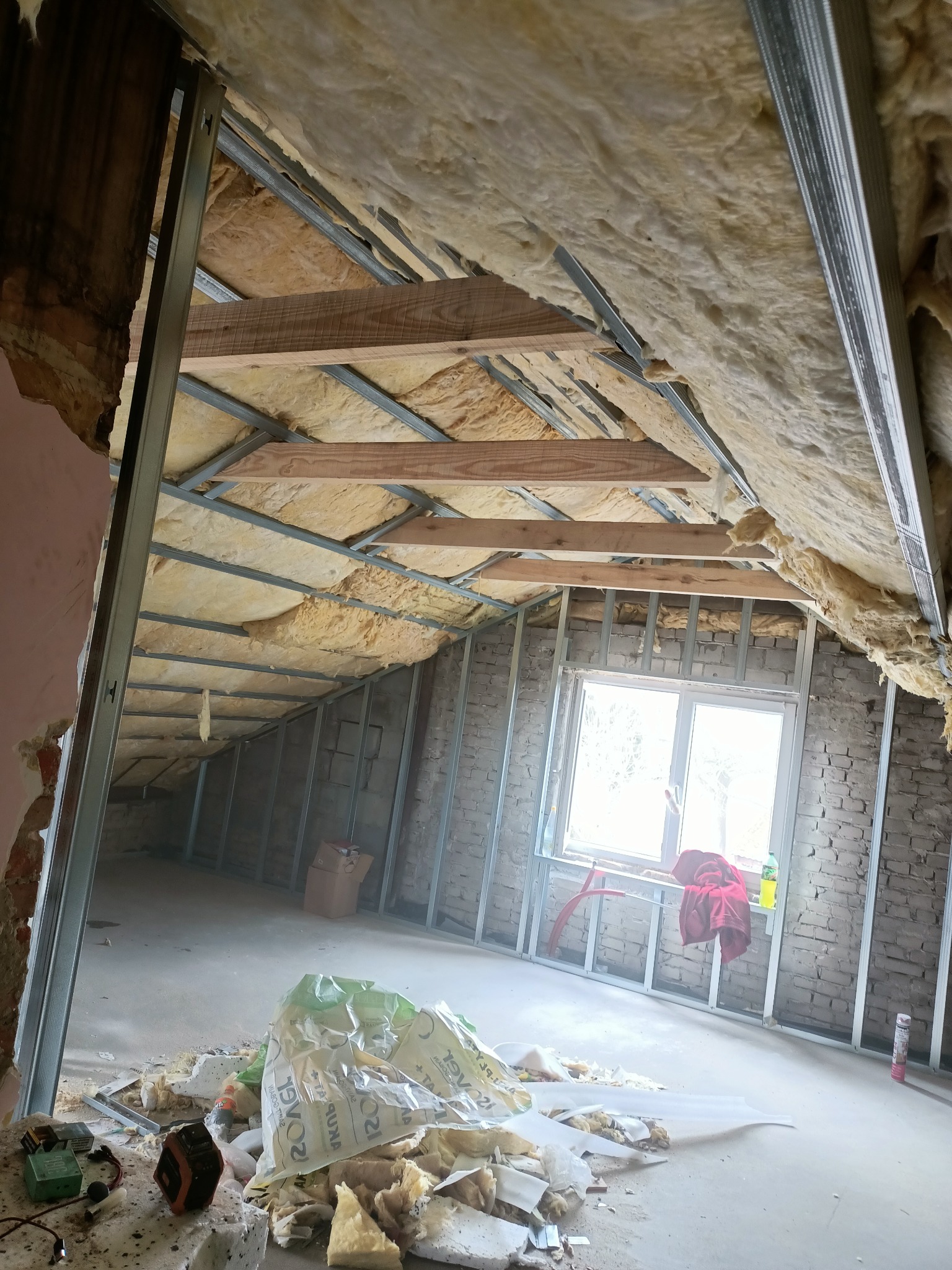 Poddasze w trakcie ocieplania wełną mineralną, widoczne metalowe profile konstrukcyjne, fragment muru z oknem, na podłodze resztki materiałów budowlanych oraz torba z wełną.