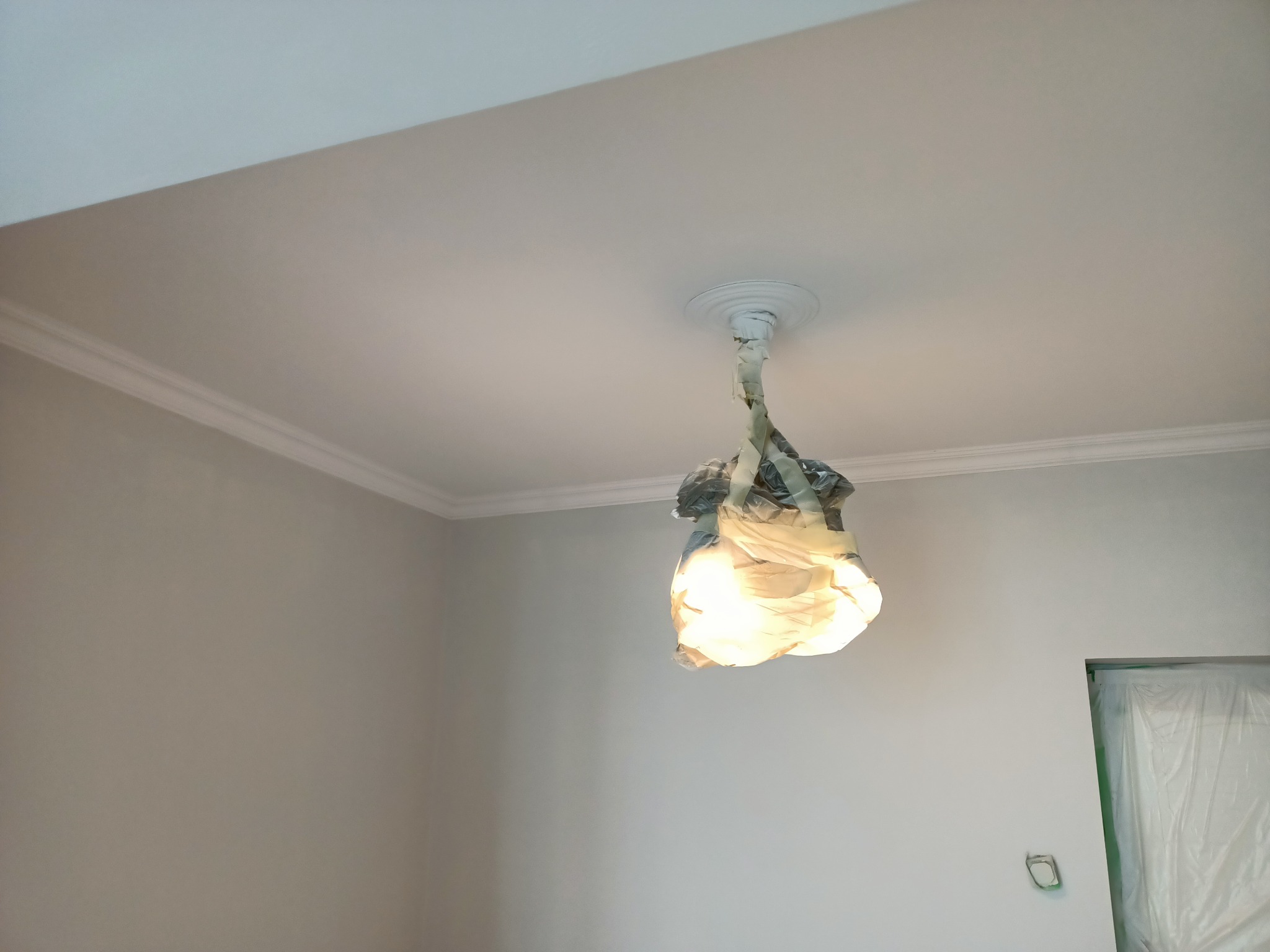 Pokój po malowaniu, widoczna świeżo pomalowana szara ściana z białym ozdobnym gzymsem pod sufitem, lampa zabezpieczona folią malarską, drzwi również zabezpieczone folią.