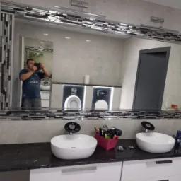 Nowoczesna łazienka z dwoma umywalkami nablatowymi, czarnym blatem i dużym lustrem w mozaikowej ramie, w odbiciu widoczny fotograf robiący zdjęcie.