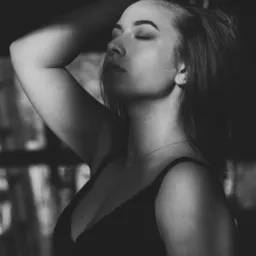 Portret kobiety w bieliźnie, z zamkniętymi oczami, delikatnie oświetlonej smugą światła na twarzy, w czerni i bieli, z kolczykiem w nosie i delikatnym łańcuszkiem na szyi, z ręką uniesioną do włosów.