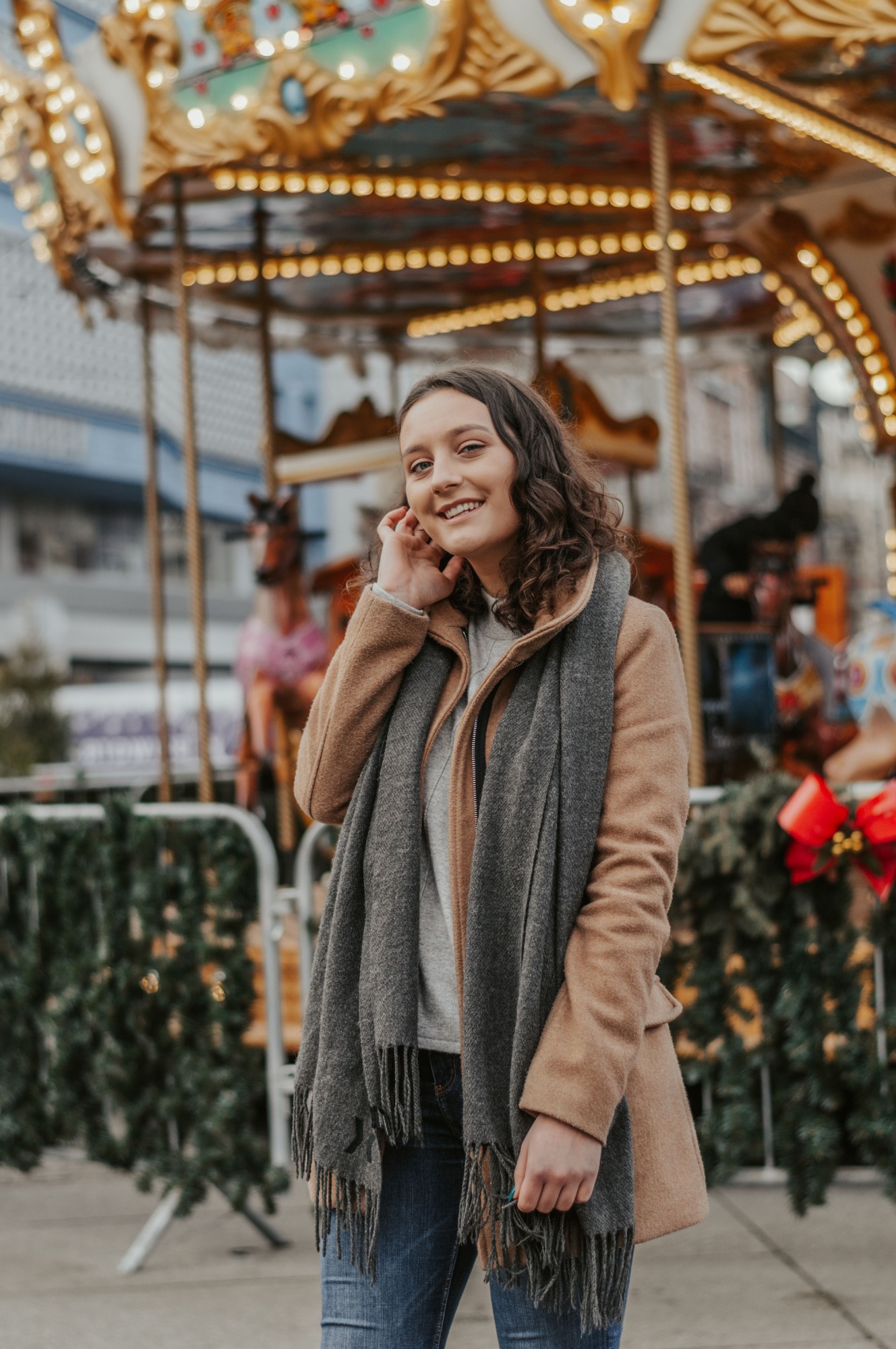 Uśmiechnięta młoda kobieta w płaszczu i szarym szalu pozuje na tle karuzeli z konikami, trzymając dłoń przy twarzy.