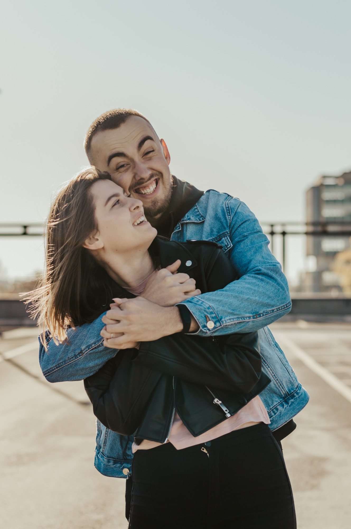 Uśmiechnięta para w objęciach na tle miejskiego parkingu, mężczyzna w jeansowej kurtce obejmuje kobietę w skórzanej kurtce, oboje patrzą w lewo.