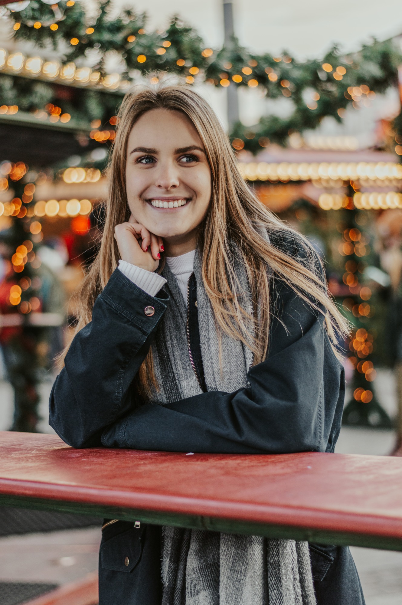 Portret uśmiechniętej kobiety w Katowicach, opierającej się o czerwony stół na tle świątecznych dekoracji i lampek bokeh.