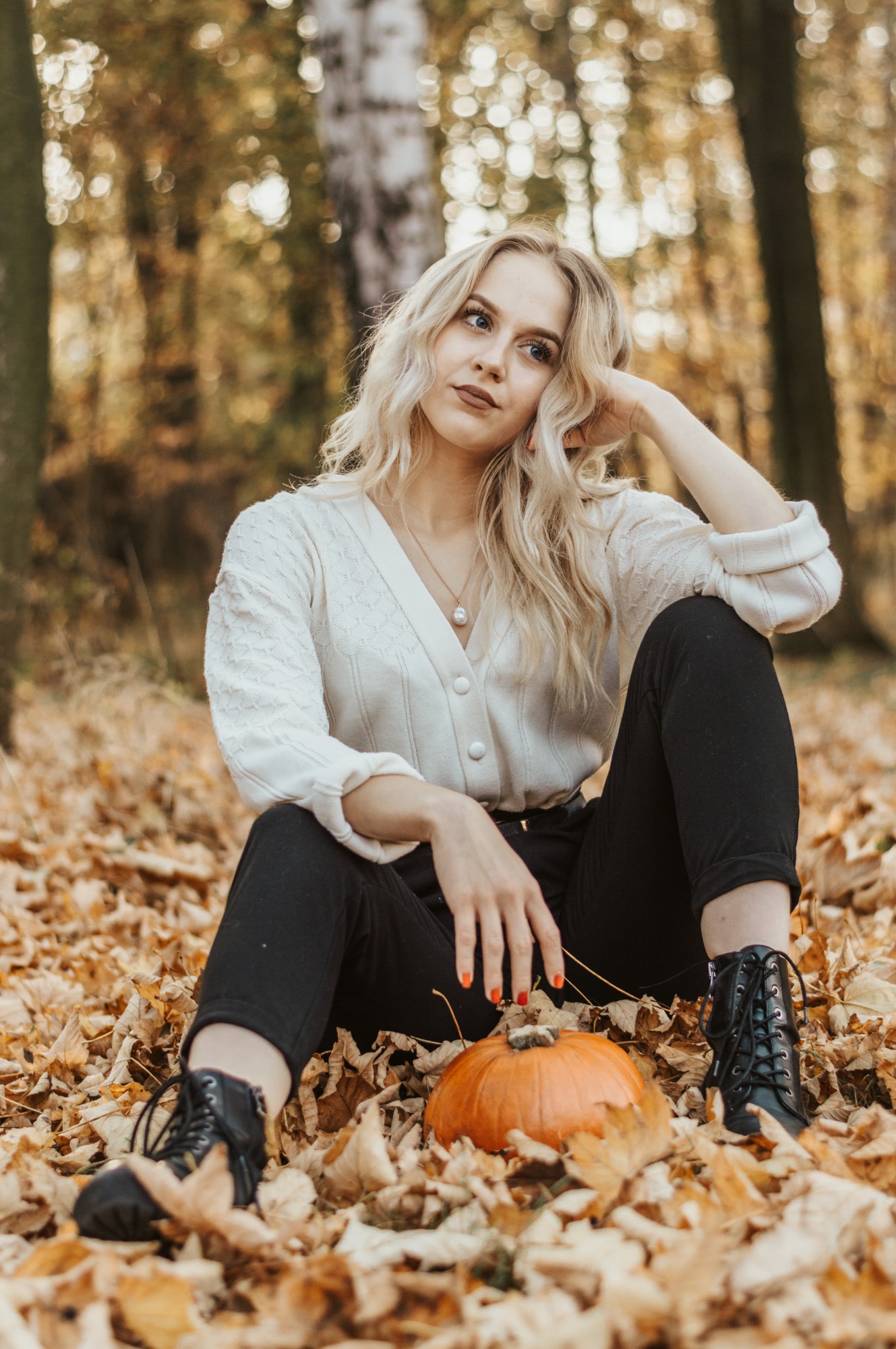 Kobieta w kremowym swetrze i czarnych spodniach siedzi na liściach w jesiennym lesie, obok niej leży mała dynia.