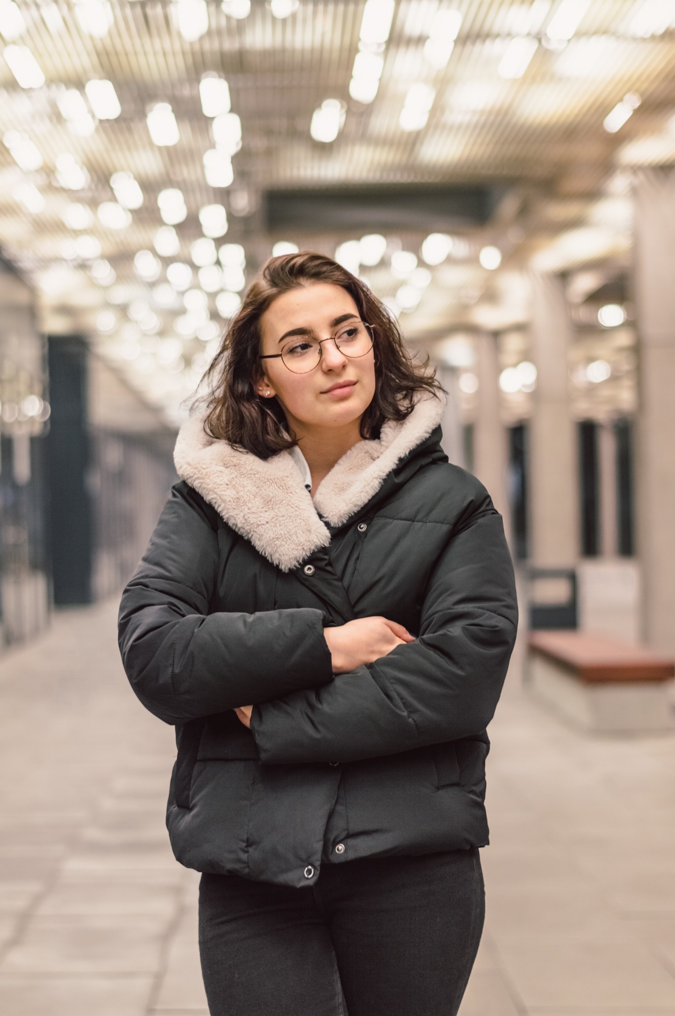 Kobieta w czarnej kurtce zimowej z futrzanym kołnierzem i okularach, z założonymi rękami, spogląda w bok na tle rozmytych świateł i filarów wewnątrz budynku.