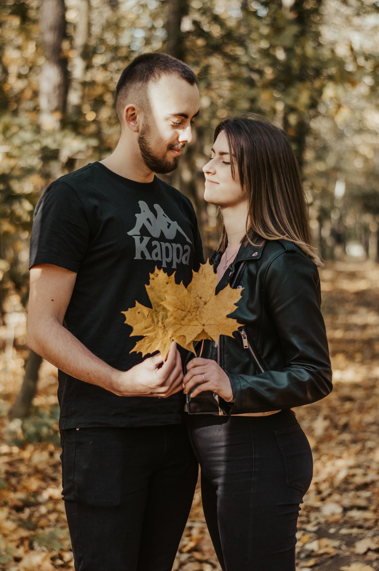 Zakochana para w jesiennym lesie, trzymająca żółte liście klonu, skierowana ku sobie z uśmiechem.