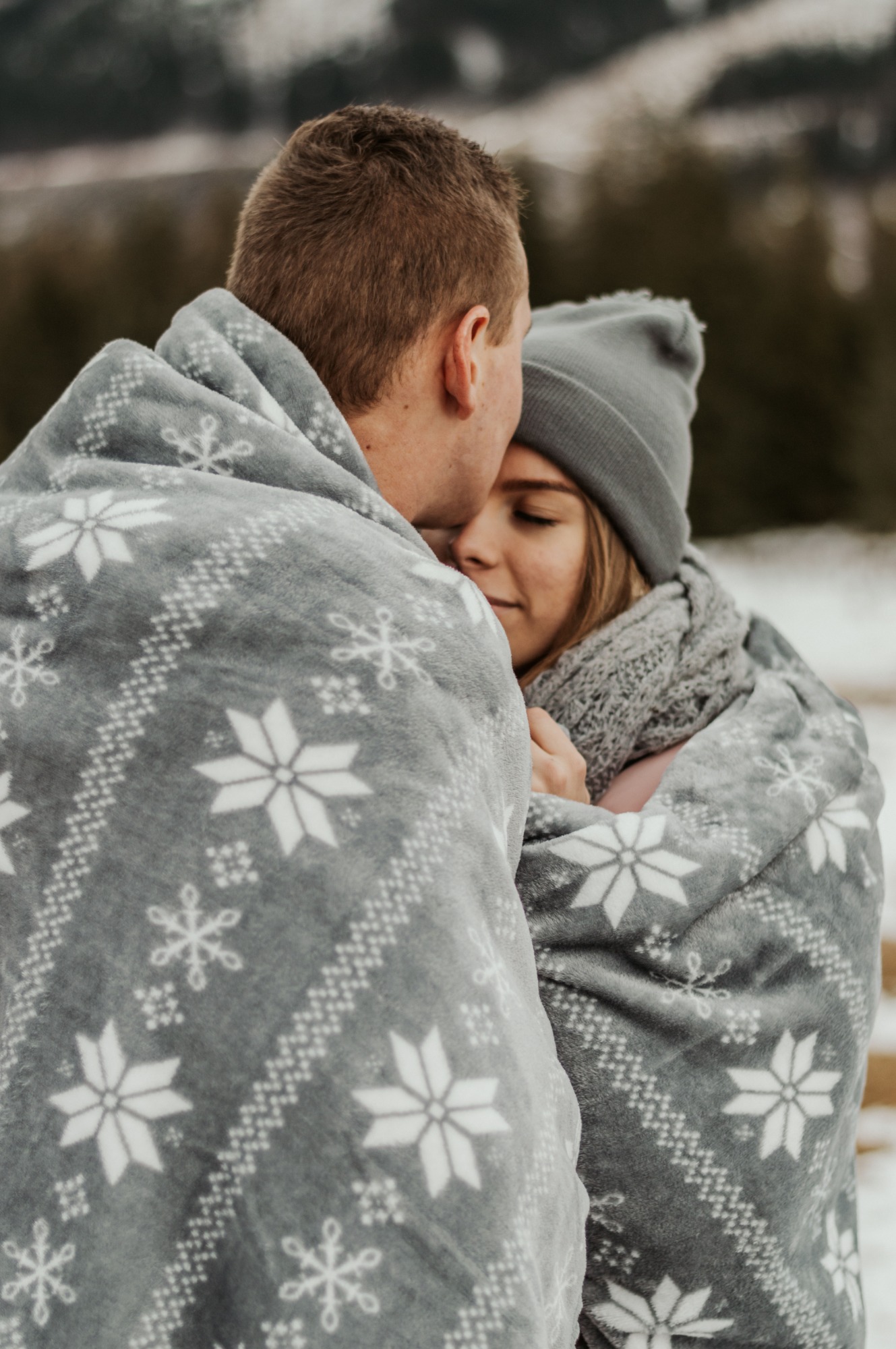 Młoda para otulona szarym kocem w białe śnieżynki, mężczyzna całuje kobietę w czapce w głowę, zimowy plener w tle.