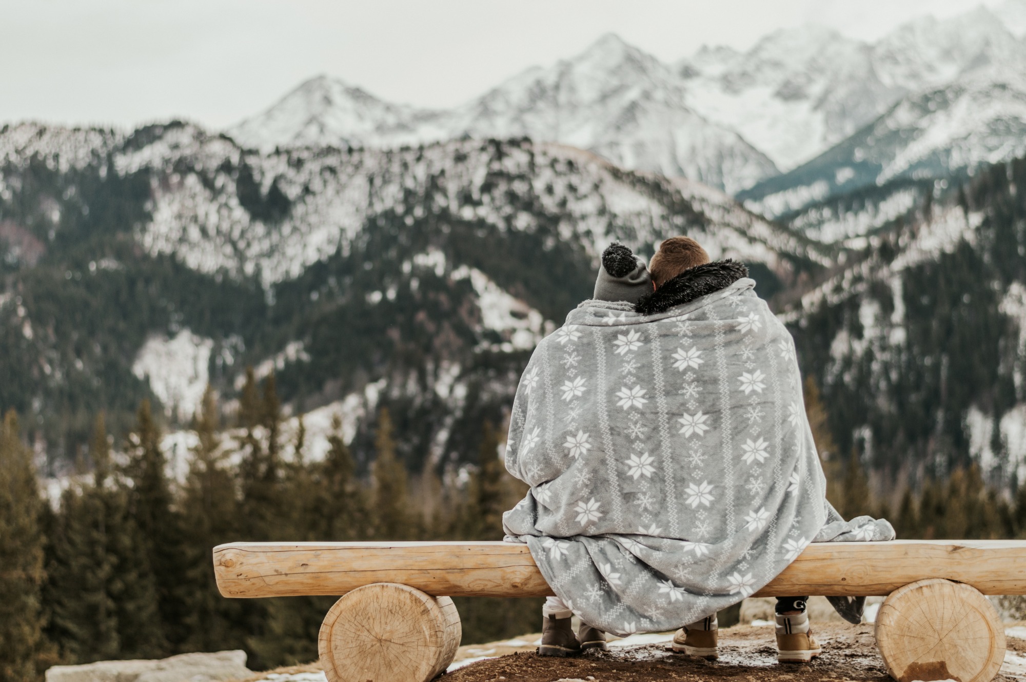 Para siedzi na drewnianej ławce, owinięta szarym kocem w białe śnieżynki, podziwiając górski krajobraz z zalesionymi zboczami i ośnieżonymi szczytami w tle.