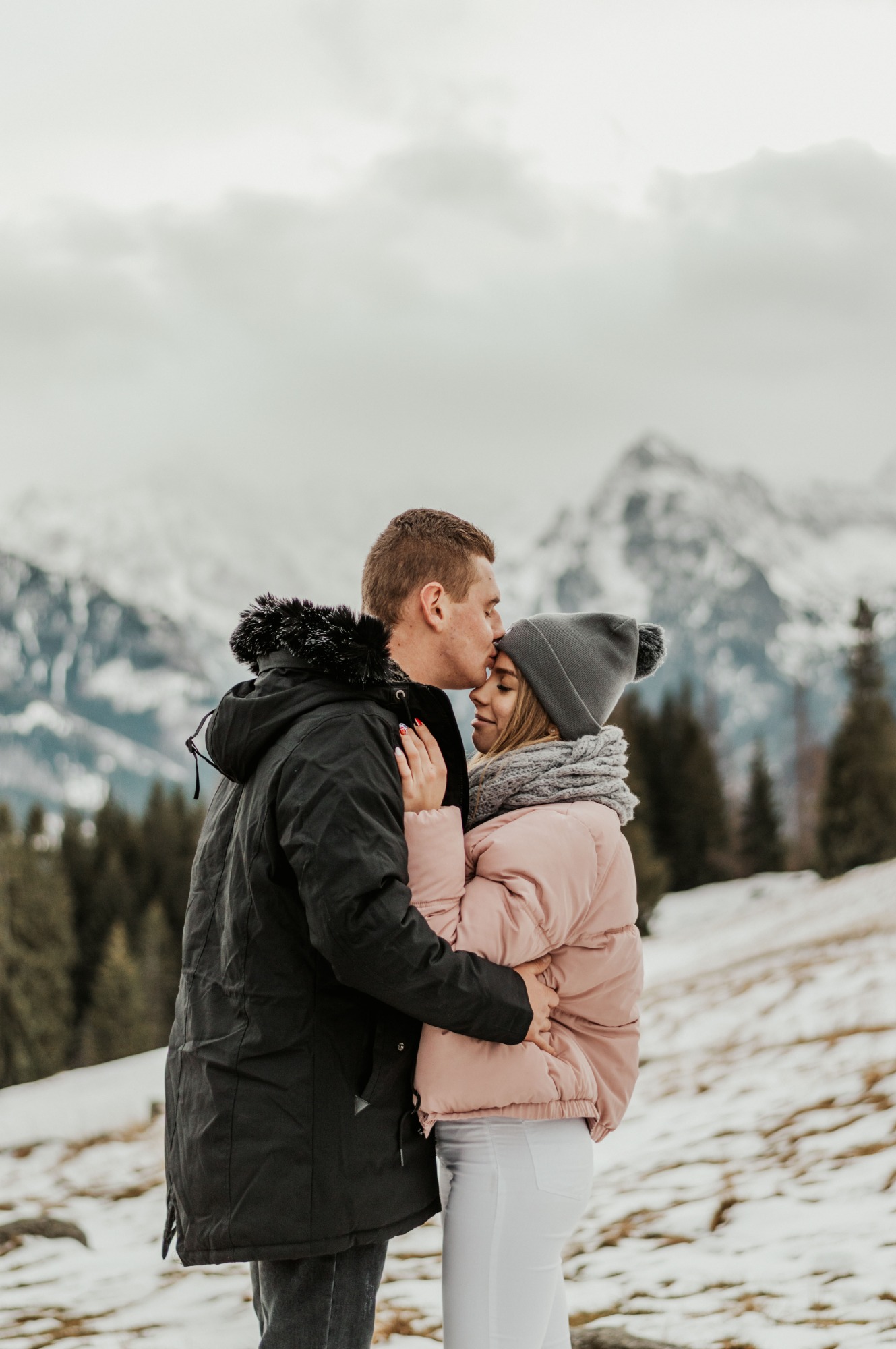 Mężczyzna całuje kobietę w czoło na tle gór; kobieta w różowej kurtce i szarym szaliku, mężczyzna w czarnej kurtce z futrzanym kapturem.