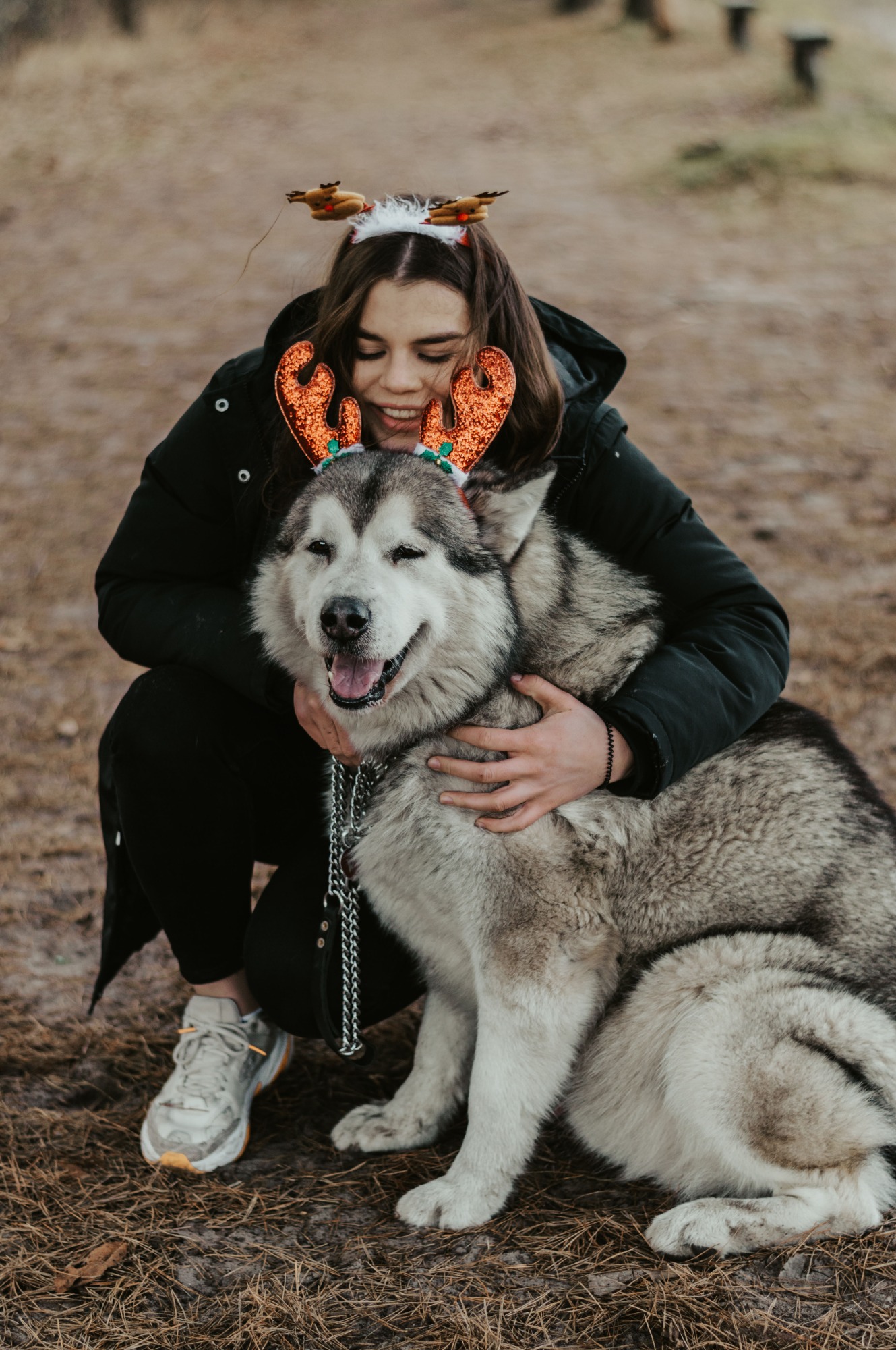 Uśmiechnięta kobieta w czarnej kurtce i reniferowych rogach obejmuje dużego psa rasy Malamut, również z rogami renifera, na tle ściółki leśnej.