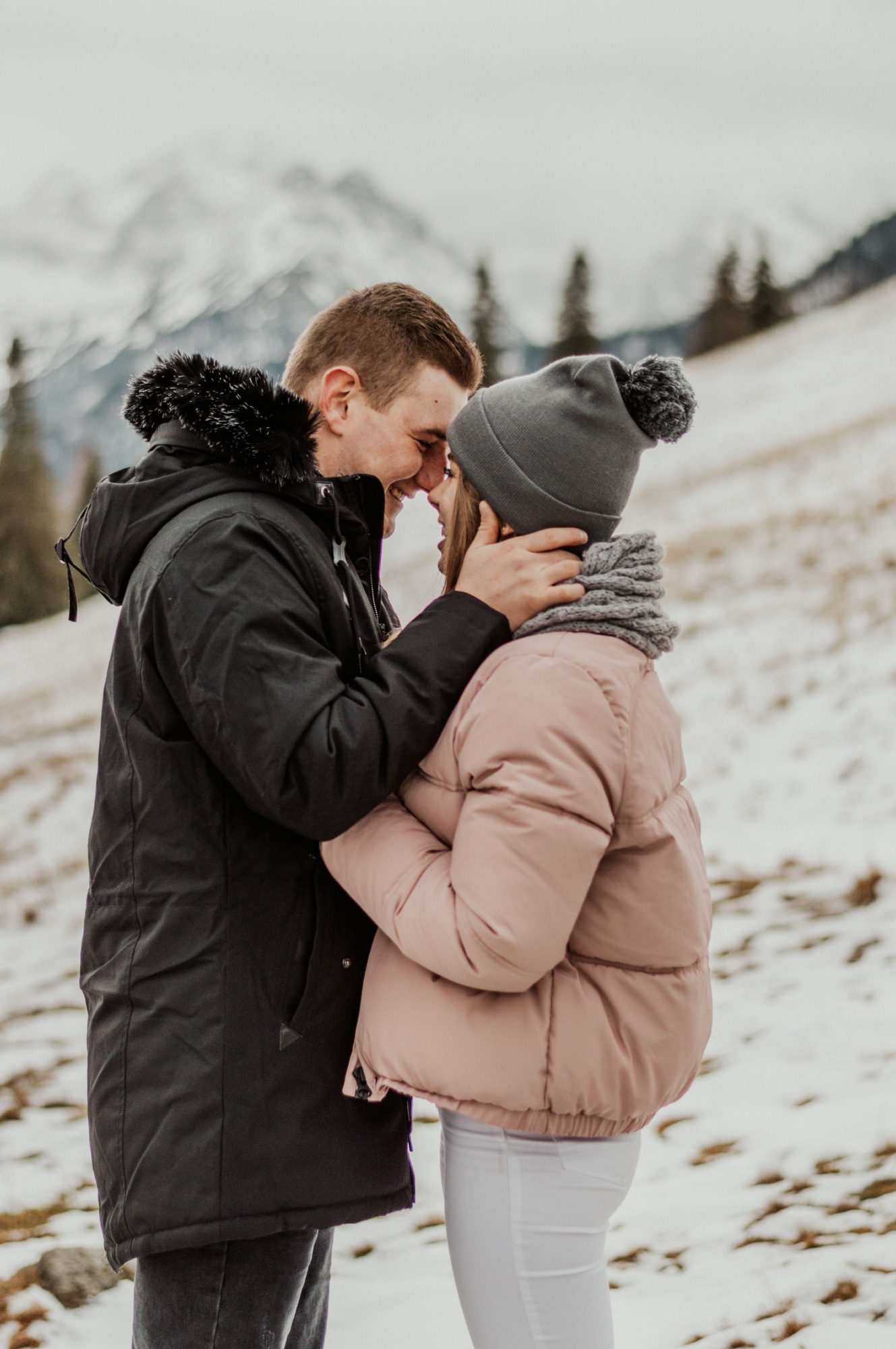 Zakochana para w zimowej scenerii; mężczyzna w czarnej kurtce z futrzanym kapturem obejmuje kobietę w różowej kurtce i szarej czapce na tle ośnieżonych gór.
