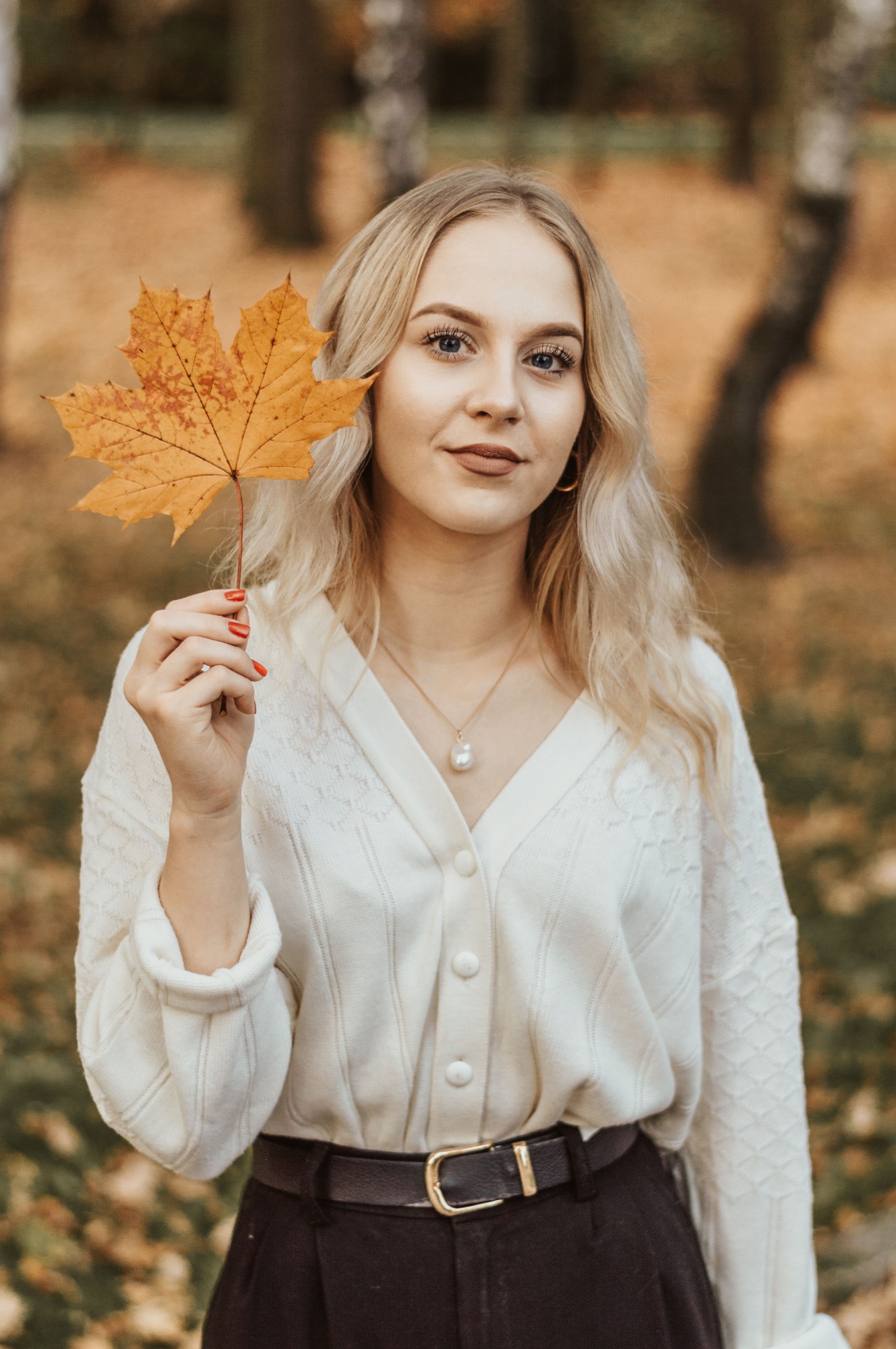 Portret młodej kobiety z blond włosami, trzymającej pomarańczowy liść klonu na tle jesiennego parku.